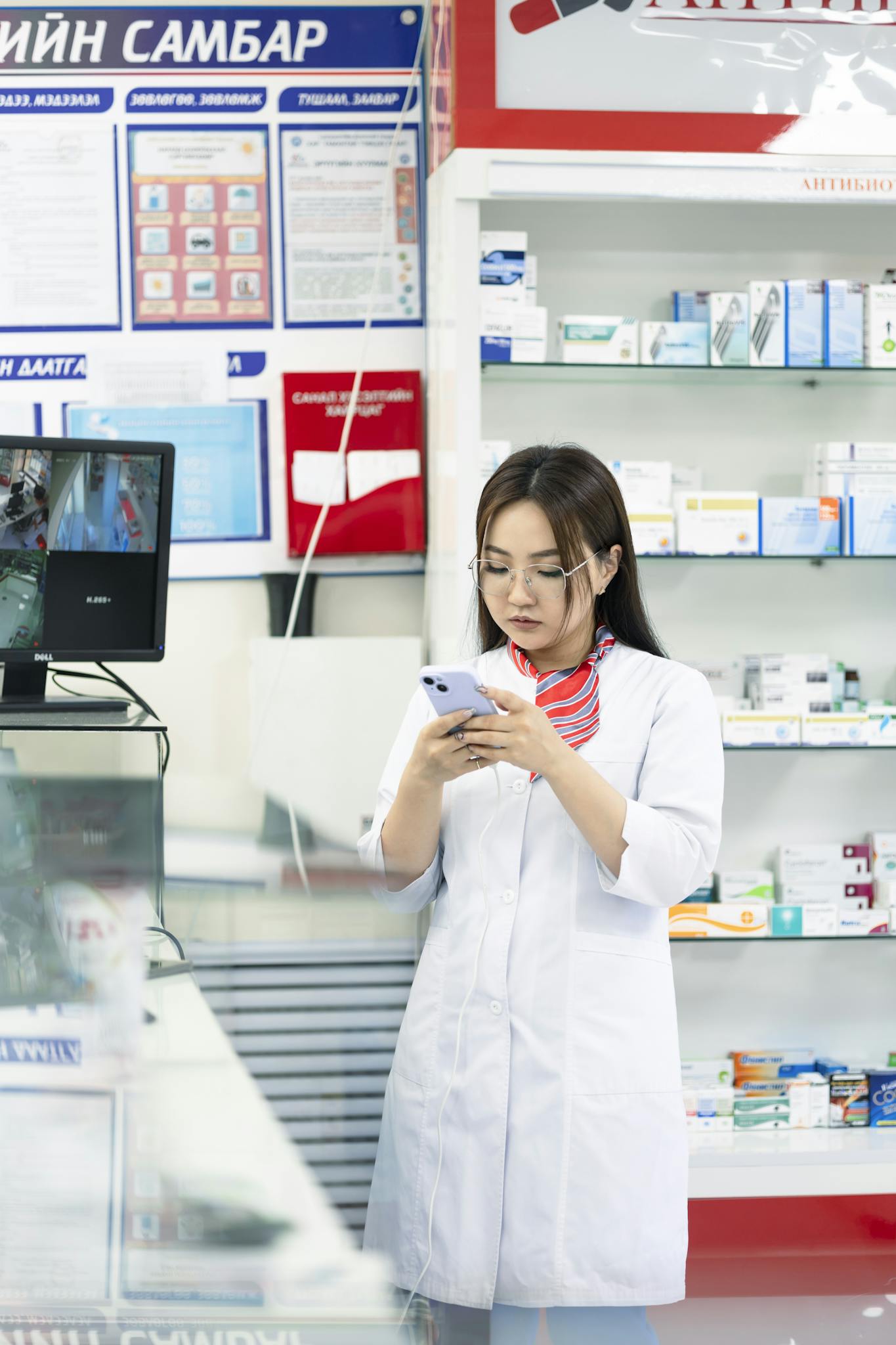 A female pharmacist in a lab coat using a smartphone in a pharmacy, surrounded by medicine shelves.