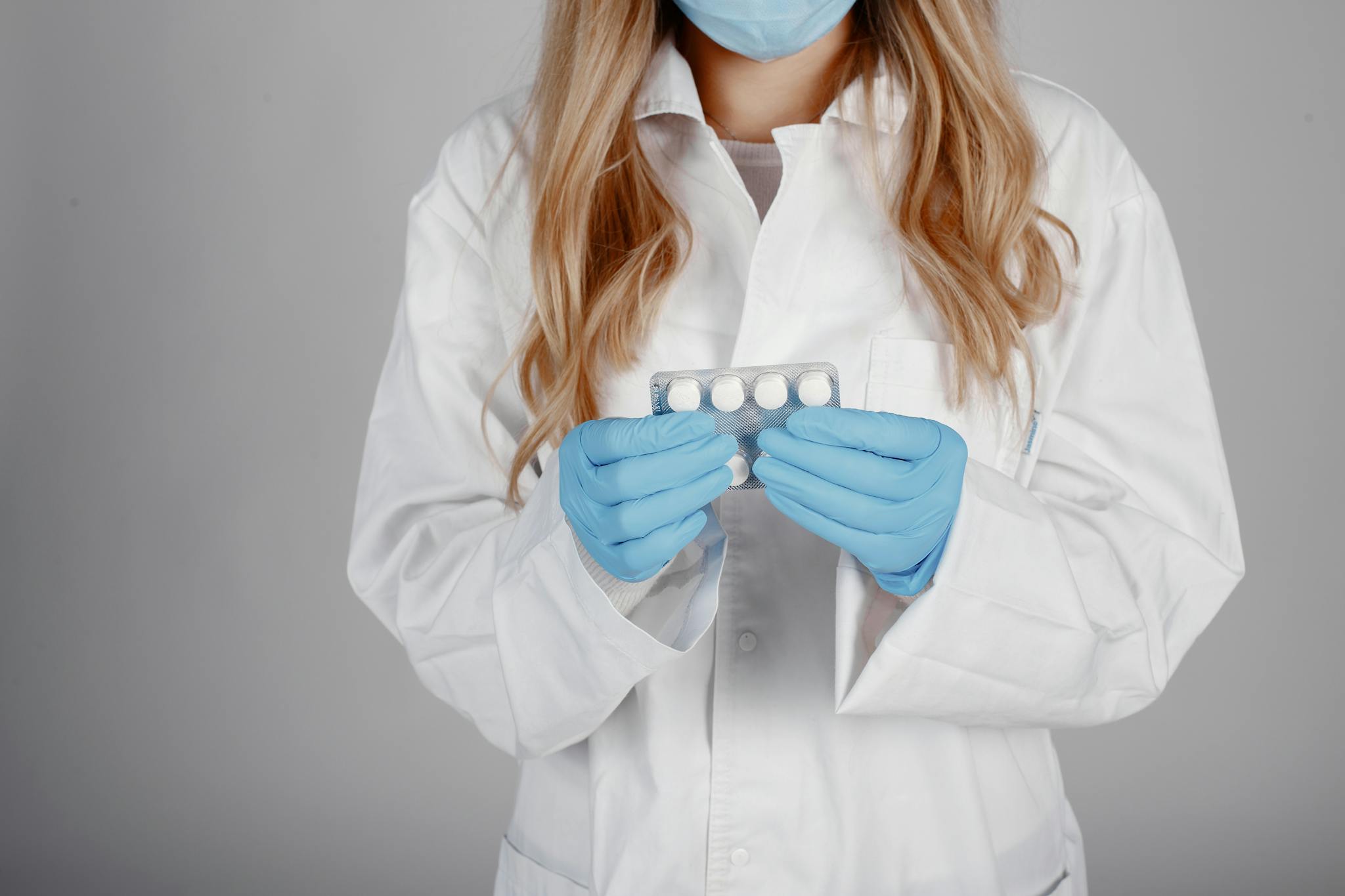 Healthcare professional in white coat and mask holding medication with gloved hands.